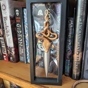 May include: A gold-coloured snake dagger with a silver blade is displayed in a black shadow box on a bookshelf. The dagger is surrounded by books with titles like "Gideon the Ninth" and "An Enchantment of Ravens".