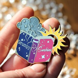 May include: A gold and silver enamel pin featuring two closed books with the words "Grumpy" and "Sunshine" on the covers. The books are surrounded by a cloud and a sun with rays.