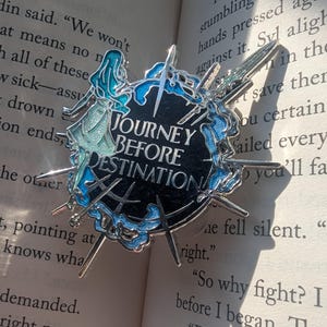 May include: A silver enamel pin with a black background and blue and white accents. The pin features a circle with the text "Journey Before Destination" in silver lettering. The circle is surrounded by six silver swords.
