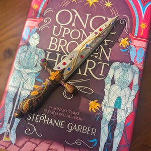 May include: A book titled "Once Upon a Broken Heart" by Stephanie Garber, a #1 Sunday Times bestselling author. The book cover features a sword with a floral design and colorful gems, two knights in armor, and a red, pink, and blue background.