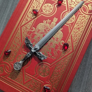 May include: A silver sword with a winged angel figure and a star-shaped pommel, resting on a red book with gold ornate designs and red gemstones scattered around it.