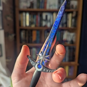 Puede incluir: Una pequeña daga con mango negro y un diseño de media luna y estrella plateadas. La hoja es de un azul profundo con un patrón de galaxia.