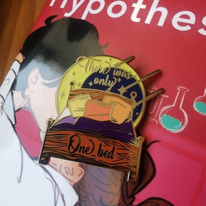 Puede incluir: Pin de esmalte dorado con una cama, almohadas y una luna con estrellas. El texto en la luna dice "There was only" y el texto en la cama dice "One bed".
