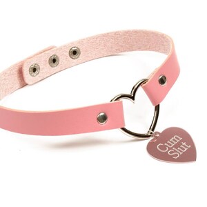 K&ouml;nnte beinhalten: Eine rosa Choker-Halskette mit einem herzf&ouml;rmigen Anh&auml;nger. Die Halskette hat ein rosa Lederband mit silberfarbener Hardware und einen herzf&ouml;rmigen Metallring. Der Anh&auml;nger ist mit den Worten "Cum Slut" graviert.