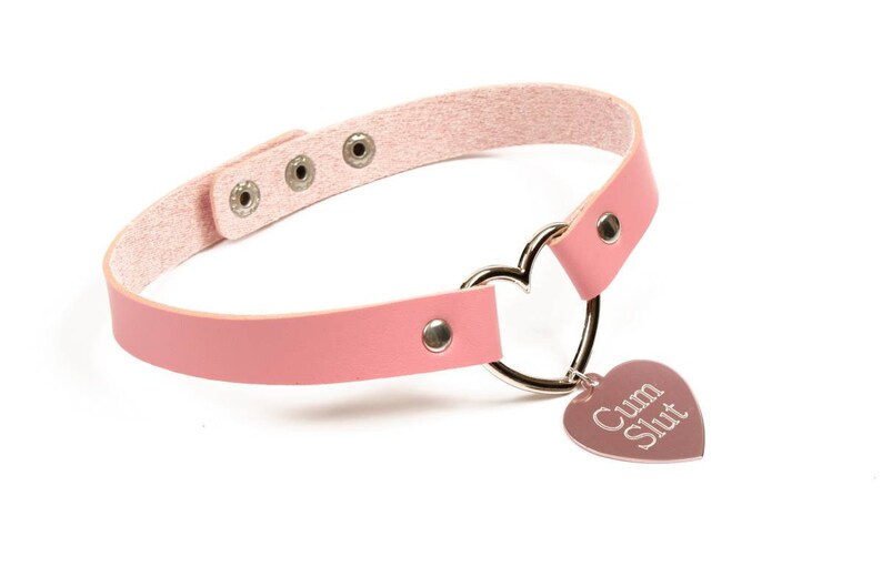K&ouml;nnte beinhalten: Eine rosa Choker-Halskette mit einem herzf&ouml;rmigen Anh&auml;nger. Die Halskette hat ein rosa Lederband mit silberfarbener Hardware und einen herzf&ouml;rmigen Metallring. Der Anh&auml;nger ist mit den Worten "Cum Slut" graviert.