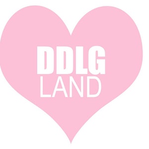 K&ouml;nnte beinhalten: Ein rosa Herz mit dem wei&szlig;en Schriftzug "DDLG LAND". Das Herz hat eine einfarbige, pastellrosa Farbe, und der Text ist zentriert innerhalb der Herzform.