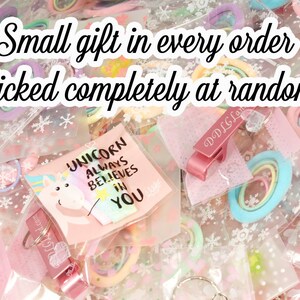 K&ouml;nnte beinhalten: Eine Auswahl an kleinen Geschenken, darunter Haargummis, Schl&uuml;sselanh&auml;nger und Karten, verpackt in transparenten Plastikt&uuml;ten. Der Text "Small gift in every order, Picked completely at random" ist zu sehen. Eine Karte mit einer Einhorn-Illustration ist ebenfalls sichtbar.