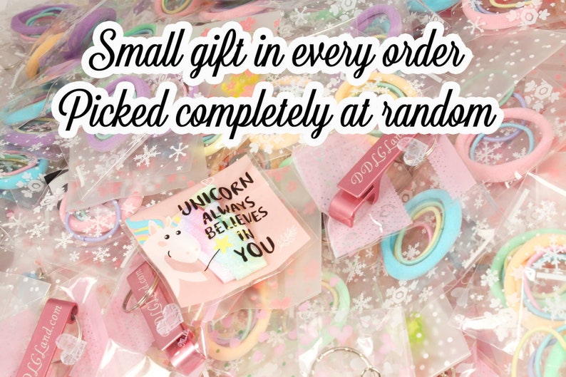 K&ouml;nnte beinhalten: Eine Auswahl an kleinen Geschenken, darunter Haargummis, Schl&uuml;sselanh&auml;nger und Karten, verpackt in transparenten Plastikt&uuml;ten. Der Text "Small gift in every order, Picked completely at random" ist zu sehen. Eine Karte mit einer Einhorn-Illustration ist ebenfalls sichtbar.