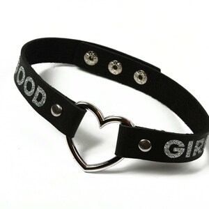 Puede incluir: Gargantilla negra con texto plateado brillante que dice "GOOD GIRL". La gargantilla presenta un colgante plateado en forma de coraz&oacute;n y cierres de presi&oacute;n plateados. El accesorio est&aacute; dise&ntilde;ado para usarse alrededor del cuello.