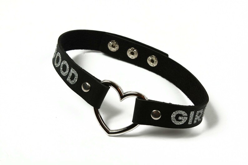 Puede incluir: Gargantilla negra con texto plateado brillante que dice "GOOD GIRL". La gargantilla presenta un colgante plateado en forma de coraz&oacute;n y cierres de presi&oacute;n plateados. El accesorio est&aacute; dise&ntilde;ado para usarse alrededor del cuello.