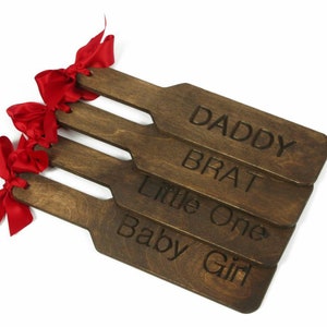 Puede incluir: Cuatro palas de madera con lazos rojos. Las palas están grabadas con las palabras "Daddy", "Brat", "Little One" y "Baby Girl".