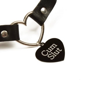 K&ouml;nnte beinhalten: Schwarzes Lederhalsband mit einem silbernen herzf&ouml;rmigen Anh&auml;nger und einem schwarzen herzf&ouml;rmigen Charm. Der Charm hat die Worte "Cum Slut" in silbernen Buchstaben. Das Halsband hat einen silbernen Ring und einen silbernen Niet.