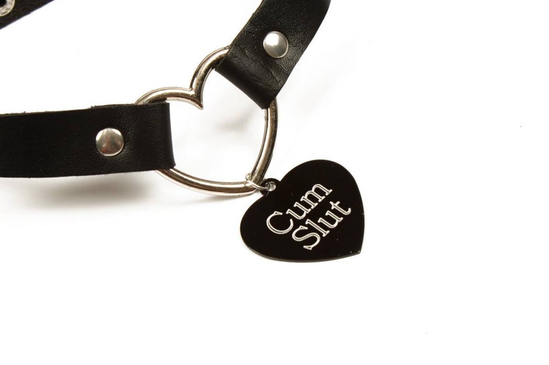 K&ouml;nnte beinhalten: Schwarzes Lederhalsband mit einem silbernen herzf&ouml;rmigen Anh&auml;nger und einem schwarzen herzf&ouml;rmigen Charm. Der Charm hat die Worte "Cum Slut" in silbernen Buchstaben. Das Halsband hat einen silbernen Ring und einen silbernen Niet.