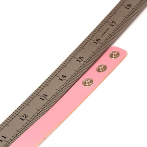 K&ouml;nnte beinhalten: Ein rosa Lederarmband mit drei silbernen Druckkn&ouml;pfen wird mit einem Edelstahl-Lineal gemessen. Das Lineal zeigt Messungen in Zentimetern an, wobei sich das Armband von der 33-cm-Marke bis zur 41-cm-Marke erstreckt.