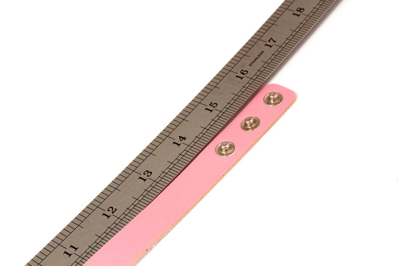 K&ouml;nnte beinhalten: Ein rosa Lederarmband mit drei silbernen Druckkn&ouml;pfen wird mit einem Edelstahl-Lineal gemessen. Das Lineal zeigt Messungen in Zentimetern an, wobei sich das Armband von der 33-cm-Marke bis zur 41-cm-Marke erstreckt.