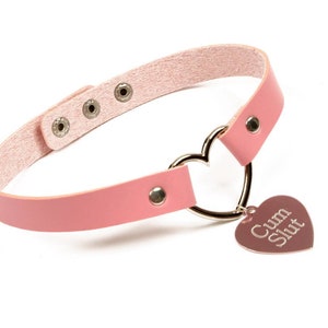 K&ouml;nnte beinhalten: Rosa Choker-Halskette mit herzf&ouml;rmigem Anh&auml;nger. Die Halskette besteht aus rosa Leder und verf&uuml;gt &uuml;ber einen silbernen herzf&ouml;rmigen Ring und einen herzf&ouml;rmigen Anh&auml;nger mit dem Text "Cum Slut".