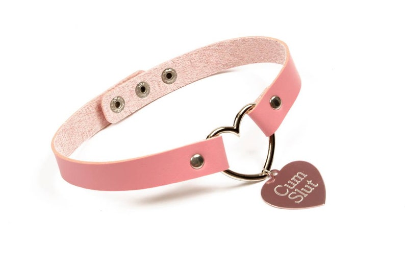 K&ouml;nnte beinhalten: Rosa Choker-Halskette mit herzf&ouml;rmigem Anh&auml;nger. Die Halskette besteht aus rosa Leder und verf&uuml;gt &uuml;ber einen silbernen herzf&ouml;rmigen Ring und einen herzf&ouml;rmigen Anh&auml;nger mit dem Text "Cum Slut".