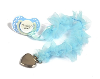 Puede incluir: Un chupete azul y blanco con el texto "Daddy's Girl" unido a una cinta de tul azul con un broche de corazón plateado.