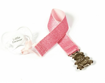 Puede incluir: Clip para chupete de color rosa con purpurina, con un dije de oso de peluche dorado y un chupete transparente con el texto "Little Forever".