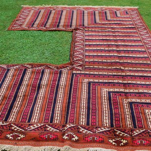 May include: An L-shaped rug with a matching smaller rug, displayed on green grass. The rugs feature a striped pattern in shades of brown, red, and cream, with geometric borders and fringed edges. The design is symmetrical and traditional.