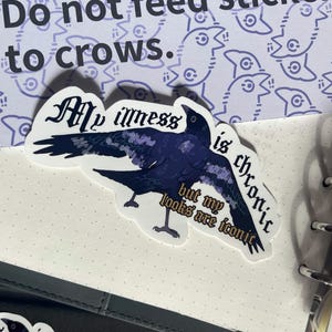 Könnte beinhalten: Ein gestanzter Aufkleber mit einem dunkelblauen Raben mit ausgebreiteten Flügeln. Der Text "My illness is chronic but my looks are iconic" ist in einer gotischen Schrift über den Vogel gedruckt. Der Aufkleber hat einen weißen Rand.