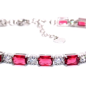 Op de afbeelding: Een zilveren armband met afwisselend rechthoekige rode en ronde heldere edelstenen. De armband is geplaatst tegen een witte achtergrond, die de glans en het ontwerp benadrukt.