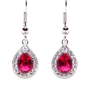 May include: A pair of silver drop earrings. Each earring has a teardrop-shaped, bright red gemstone, encircled by small, clear crystals. The earrings are suspended from a simple hook.