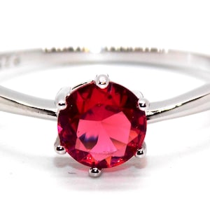 May include: A silver ring featuring a bright red gemstone. The round-cut stone is held in place by six prongs, and the band is smooth and polished. The ring is shown against a white backdrop.