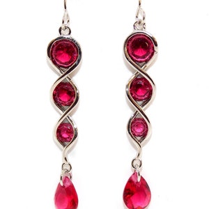 May include: Dangling earrings with a silver-tone metal design. Each earring features three round, bright pink gemstones and a teardrop-shaped pink gemstone at the bottom. The earrings hang from a silver hook.