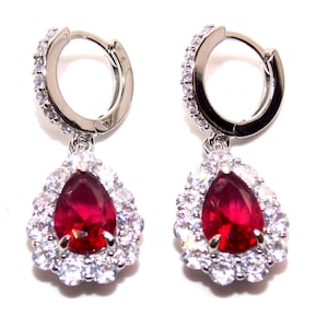 May include: A pair of silver hoop earrings. Each earring has a teardrop-shaped, deep red gemstone surrounded by a halo of smaller, clear gemstones. The top of each earring is also adorned with clear gemstones.