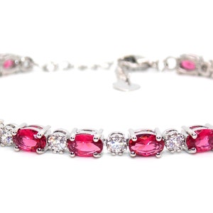 May include: A silver tennis bracelet featuring oval-shaped red gemstones and smaller clear gemstones.