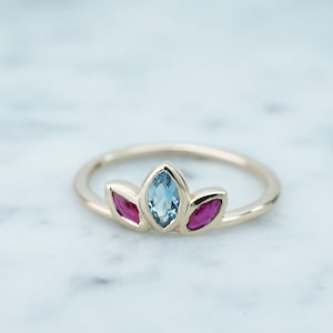 Puede incluir: Un anillo de oro con un diseño floral, con una piedra preciosa azul claro en el centro y dos piedras rojas a cada lado. El anillo está colocado sobre un fondo de mármol blanco.