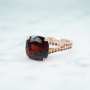 May include: A gold ring with a large, dark red gemstone set in a cushion cut. The band is accented with smaller gemstones.