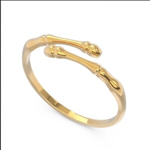 K&ouml;nnte beinhalten: Ein goldfarbener Ring mit einem einzigartigen Design, das zwei ineinander verschlungene Knochen nachahmt. Der Ring ist offen und verstellbar.