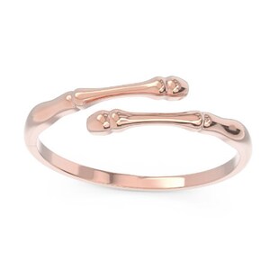 K&ouml;nnte beinhalten: Ein Ring aus Ros&eacute;gold mit einem einzigartigen Design, das zwei knochenf&ouml;rmige B&auml;nder zeigt, die sich um den Finger wickeln.