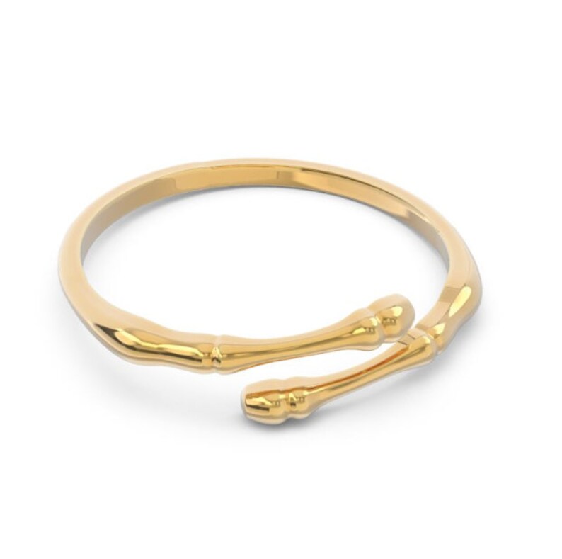 K&ouml;nnte beinhalten: Ein goldfarbener Ring mit einem einzigartigen Design aus zwei geschwungenen, abgerundeten St&auml;ben, die sich kreuzen. Der Ring ist aus glattem, poliertem Metall.