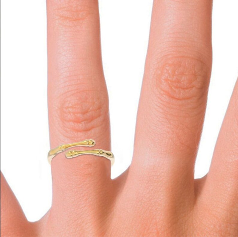 K&ouml;nnte beinhalten: Ein goldfarbener Ring mit einem einzigartigen Design, das zwei Knochen &auml;hnelt, die umeinander gewickelt sind. Der Ring wird an einem Finger getragen.
