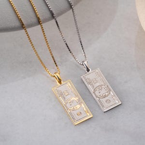 May include: Two rectangular pendant necklaces, one gold-toned and one silver-toned, each featuring a detailed design of a $100 bill. The pendants hang from delicate box chains. The gold necklace has a warm, shiny finish, while the silver one has a cool, metallic appearance.