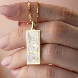 Könnte beinhalten: Eine goldfarbene Halskette mit Anhänger, die einen rechteckigen Anhänger hat, der einer 100-Dollar-Note nachempfunden ist. Der Anhänger zeigt die Zahl "100" und ein Porträt. Die Halskette wird von einer Person gehalten.