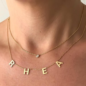 May include: A gold chain necklace with a small clear stone charm and a pendant with the letters "RHEA" hanging from it.