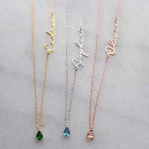 Puede incluir: Tres delicados collares con nombres en escritura cursiva. Uno es dorado con una gema verde en forma de lágrima, otro es plateado con una gema azul, y el tercero es de oro rosa con una gema melocotón. Los nombres son "Valeria", "Sophia" y "Olivia".
