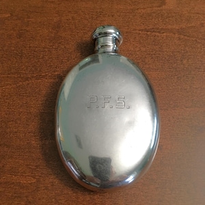 Puede incluir: Una petaca de metal plateado pulido con forma ovalada y un tapón de rosca redondeado. La superficie frontal de la petaca presenta las letras grabadas P.F.S. centradas en el medio del cuerpo, mostrando un acabado liso y reflectante.