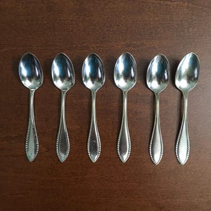 Puede incluir: Seis cucharas plateadas dispuestas sobre una superficie de madera marrón oscuro. Las cucharas tienen un diseño clásico con un cuenco en forma de lágrima y un patrón de cuentas a lo largo del mango.
