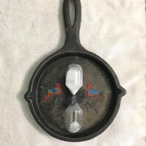 Peut inclure: Une poêle en fonte sombre et patinée avec un long manche percé d'un trou. La surface intérieure présente deux petits oiseaux peints en rouge et bleu, avec un sablier en verre transparent contenant du sable blanc placé au centre.