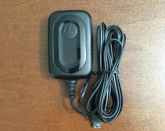 Pode incluir: Um carregador de parede Motorola preto com um adaptador de energia retangular com um painel central brilhante exibindo o logotipo da Motorola, conectado a um longo cabo de carregamento preto enrolado com um conector micro-USB padrão.