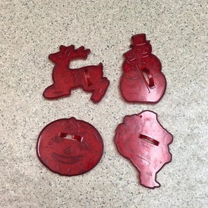 May include: A set of four red plastic cookie cutters in the shapes of a reindeer, a snowman, a pumpkin, and a turkey. Each cutter features a small handle on the back for gripping. The turkey cutter has the text MADE IN U.S.A. PAT PEND. embossed.