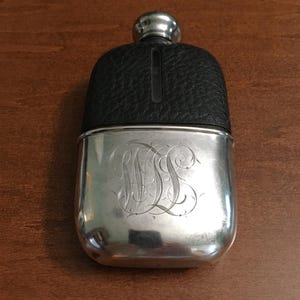 Puede incluir: Una petaca de metal plateado con una cubierta de cuero negro texturizado en la parte superior y una sección inferior de plata pulida grabada con un monograma ornamentado y en espiral. La petaca incluye un tapón plateado redondeado en la parte superior.