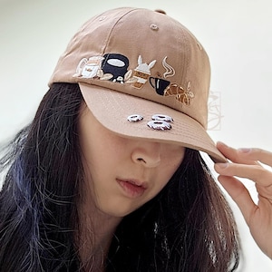 May include: A tan baseball cap featuring embroidered cartoon motifs, such as a coffee cup and a rabbit. The cap has a curved peak and a traditional six-panel construction. The neutral colour makes it suitable for various outfits and occasions.