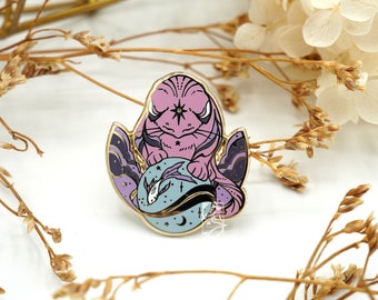 May include: A gold-toned enamel pin featuring a purple and blue creature with a star on its forehead. The creature is curled up in a ball, with its tail wrapped around its body. The pin has a black and white background with stars and a crescent moon.
