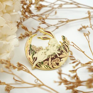 May include: Gold-tone enamel pin featuring a white rabbit hopping through a moon-shaped garden with brown, green, and gold foliage.
